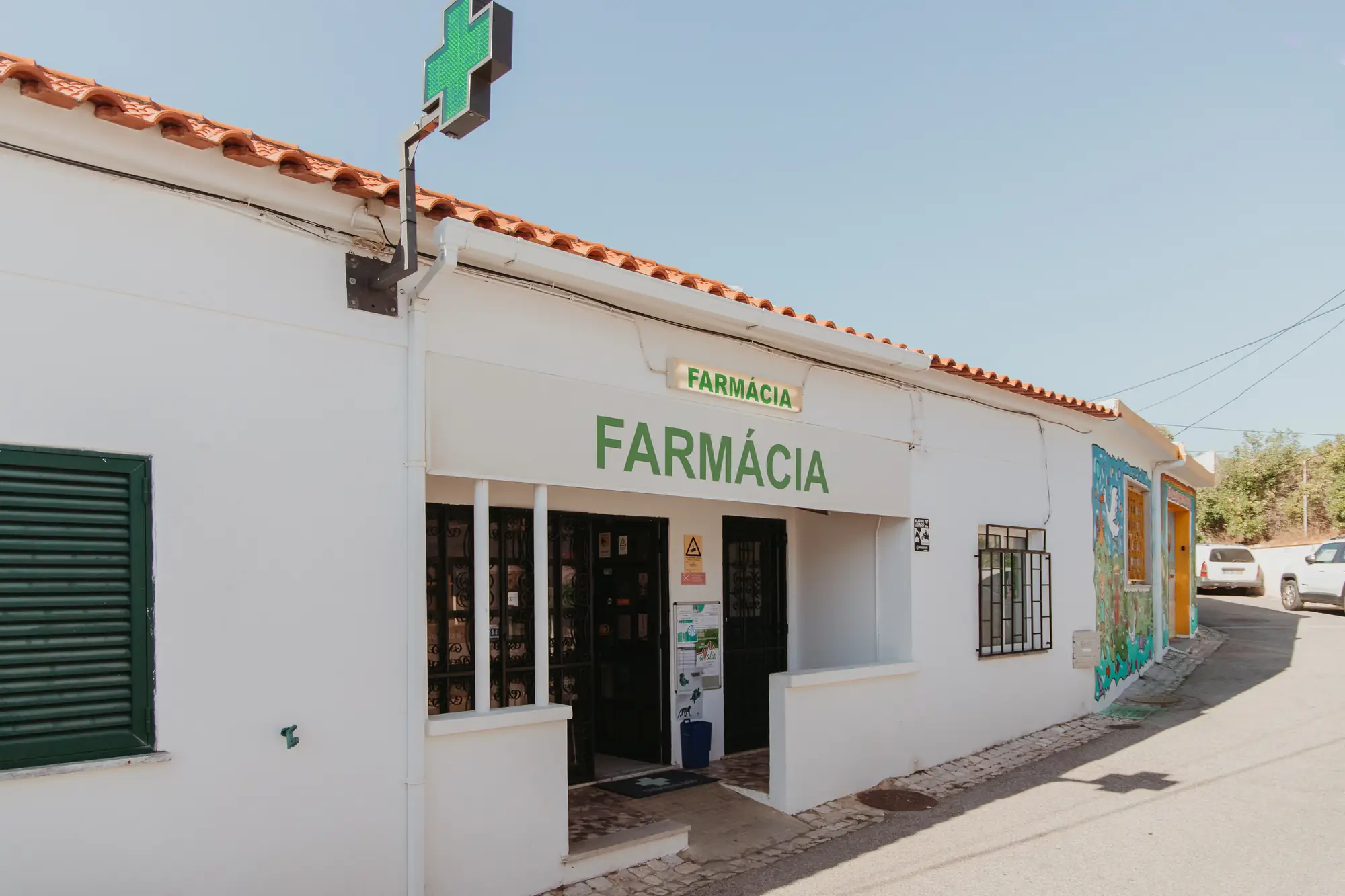 Fachada branca da Farmácia Ilda com a cruz verde acesa, na Mexilhoeira Grande